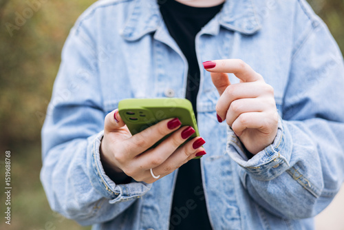 Female hands with red manicure holding green phone in denim jacket.