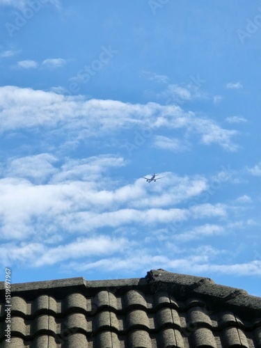 Airplane flying high in the blue sky above tiled rooftop with scattered clouds, symbolizing travel, aviation, suburban life, freedom, and sunny weather