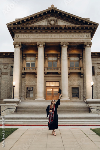Graduation photo from the University of Redlands, a girl in a robe holds her graduation hat with her hand raised to the mountain, standing in front of the university entrance.