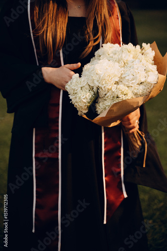 Fragment of a graduation photo shoot: a girl in a black academic gown with a burgundy ribbon holds a voluminous bouquet of white flowers wrapped in kraft paper. 