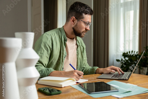 Bearded man reviewing notes on desk while working online with laptop and papers