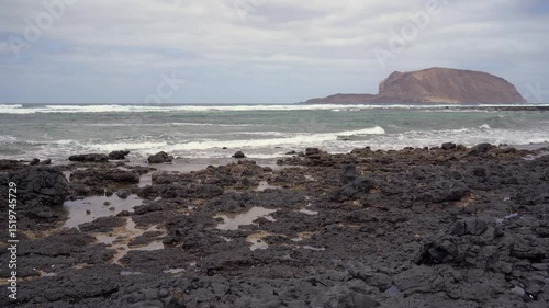 A view of La Graciosa's rocky shore under a cloudy sky, with waves crashing and a bird in flight.