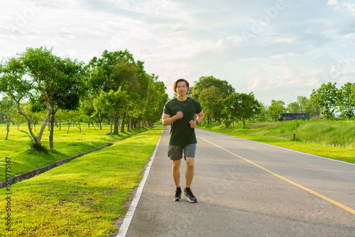 Sports man running on road for outdoor workout with warm light of sunset on freshy green background in rainy season. Jogging in nature. Healthy lifestyle.