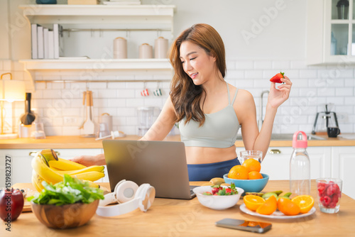 Healthy lifestyle Asian woman enjoys a peaceful weekend morning at home, relaxing after daily workout with a fresh smoothie and pure water while checking email on her laptop, embracing wellness home