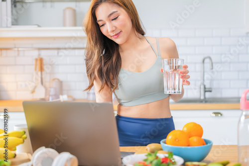 fit Asian female in sportswear enjoys a colorful fruit shake and fresh mineral water while unwinding at home in the early hours, embracing a mindful lifestyle and remote work wellness routine