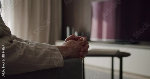 Contemplative Person Sitting on Couch with Hands Clasped Inside Cozy Home. A contemplative individual sits on a couch with hands clasped, indoors, evoking thoughts and serenity in a cozy home setting.