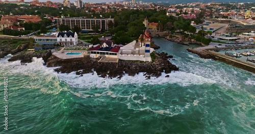 Aerial view hugging rocky coast by the sea in Cascais, Portugal with waves crashing underneath