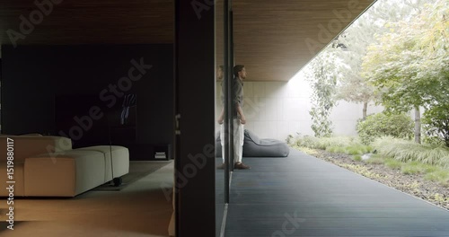 A professional man stands contemplating the serene garden view through the vast glass wall of a stylish modern home. Man Contemplating Nature from a Modern House Glass Wall
