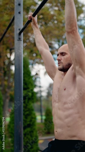 A muscular man performs pull-ups on a horizontal bar in an outdoor gym. He focuses on his form and strength, showcasing his fitness routine. Ideal for fitness, health, and workout videos.
