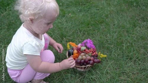 child with a basket of fruit, little girl sitting on the grass with a basket of fruit and smelling a flower