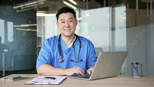Portrait smiling asian doctor in blue coat works on laptop sitting at desk at workplace in modern hospital clinic. Happy male professional medical worker physician posing in office looking at camera