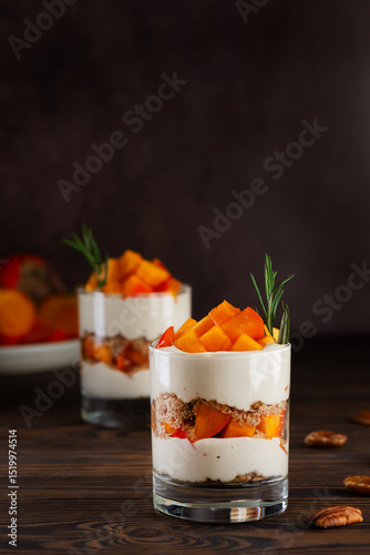 Parfait in a glasses with persimmon, rosemary, pecan, whipped cream and biscuit on a brown background. Healthy food, vegan, sugar, gluten and lactose free. Vertical orientation.