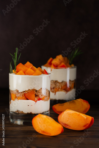 Layered dessert in a glasses with persimmon slices, rosemary, pecan, whipped cream and biscuit on a wooden table. Healthy food, vegan, sugar, gluten and lactose free. Vertical orientation.