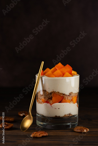 Vegan parfait in a glass with persimmon, rosemary, pecan, whipped cream and golden spoon on a brown table. Healthy food, sugar, gluten and lactose free. Vertical orientation.