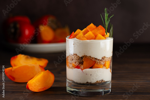 Trifle in a glass with pieces of persimmon, rosemary, pecan, whipped cream and biscuit on a brown. Healthy food, vegan, sugar, gluten and lactose free.