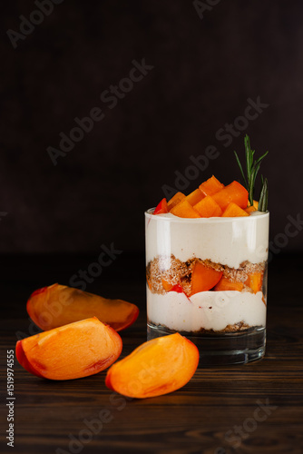 Vegan trifle in a glass with persimmon slices, rosemary, pecan, whipped cream and biscuit on a brown. Healthy food, sugar, gluten and lactose free. Vertical orientation.