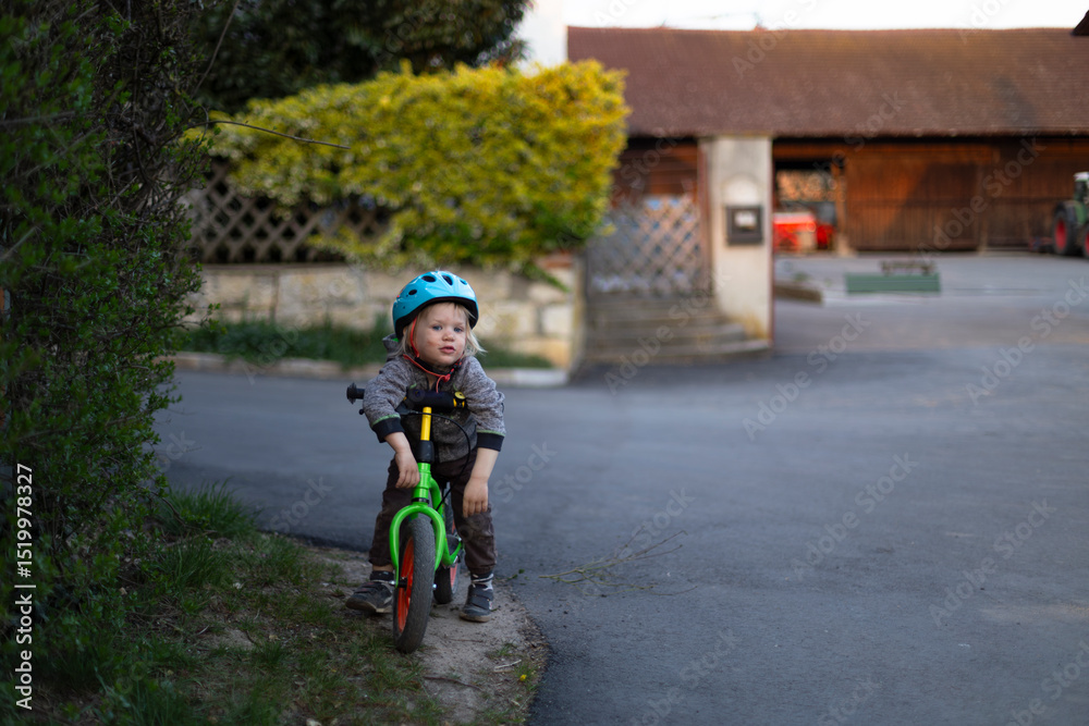Obraz premium Little tired dirty child wearing helmet riding a green balance bike in a quiet countryside village in Bavaria, Germany at sunset.