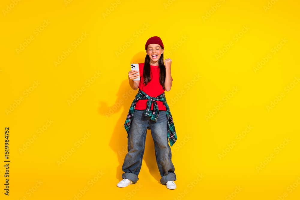 Happy young girl cheering while looking at smartphone against yellow background portraying joy and excitement Happy young girl cheering while looking at smartphone against yellow background portraying joy and excitement