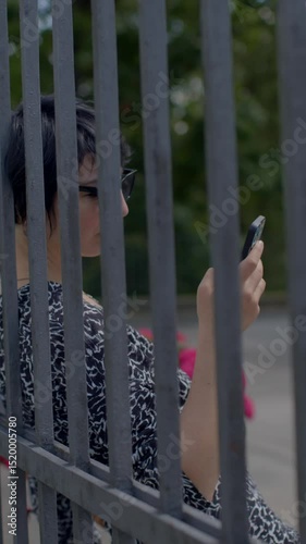 In a peaceful garden area filled with vibrant greenery and blooming flowers, a woman is comfortably seated near a sturdy fence, engrossed in her smartphone, using it during her leisure time