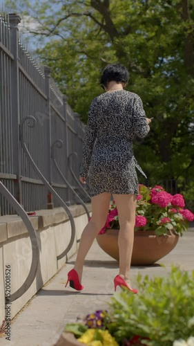 A woman casually strolls along a winding path, holding her smartphone and enjoying the beautiful scenery created by vibrant flower pots in a beautifully landscaped outdoor area