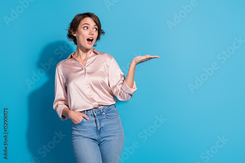 Charming young woman in a stylish satin blouse gesturing delightfully on a blue background, radiating joy and elegance
