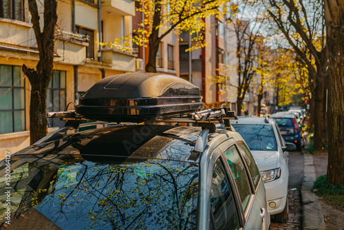 Car with rooftop cargo box parked on urban street lined with trees, warm sunset light in autumn. Concept of road trip, travel gear, exploration, ,car travel and auto gear