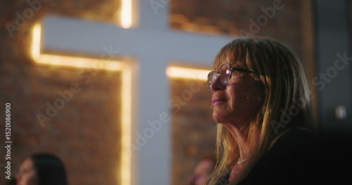 Woman with glasses looking up, emotional moment in front of illuminated cross at church during religious service, deep contemplation, faith, spiritual experience