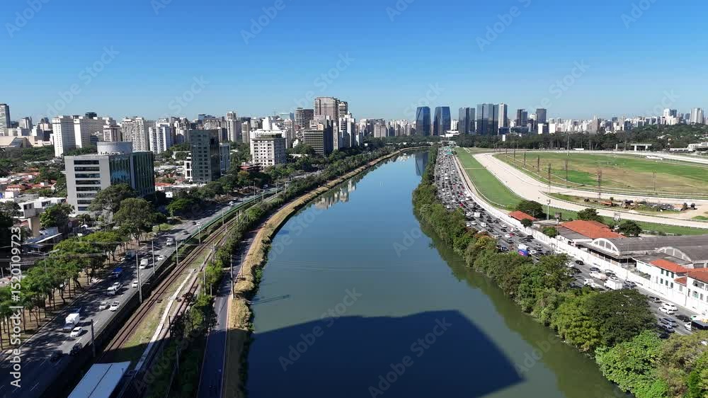 Imagem urbana mostra tráfego na Marginal Pinheiros com carros em movimento ao longo do rio e edifícios modernos ao redor