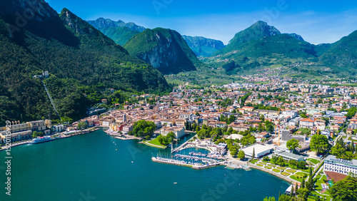 Aerial view of Riva del Garda with Torre Apponale, MAG museum, bike paths, boats and river ferries connecting towns, surrounded by mountains and clear Lake Garda water, full of tourists