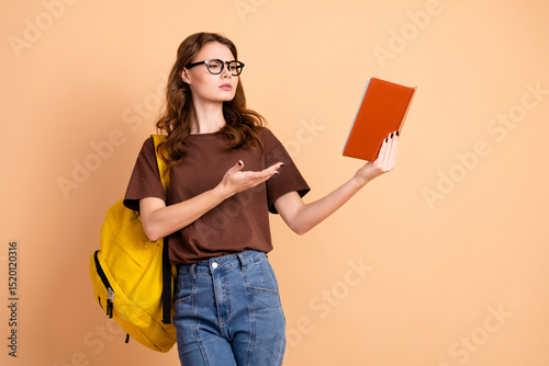 Young female student posing with orange book in casual stylish attire against beige background