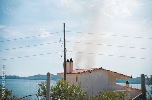 A serene summer day in Zakynthos showcases a quaint home by the sea, with smoke gently wafting from its chimney. This scene captures the essence of laid-back island life, embraced by azure waters