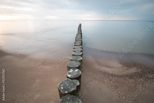 A wooden breakwater on a sandy beach is washed by blurry sea water.