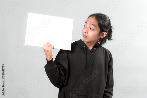 Asian man in black hoodie holding a blank white sign while looking at it with a curious expression, isolated on gray background, perfect for advertisement or message mockup.
