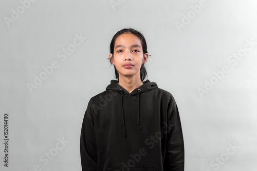 Portrait of a young Asian person standing against a plain gray background, wearing a black hoodie, looking directly at the camera with a neutral expression.