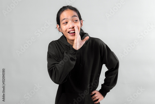 Happy young Asian man in black hoodie smiling and cupping his hand to his mouth while speaking or calling out, isolated on gray background. Concept of casual communication or greeting.