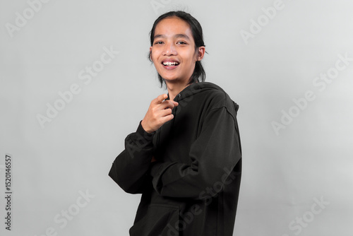 Smiling Asian man in black hoodie pointing at camera with cheerful expression, suggesting confidence, friendliness, and direct communication, on plain light gray background.