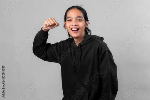 Excited Asian man in black hoodie showing cheerful expression with raised fist gesture, celebrating success or achievement, isolated on clean studio gray background.