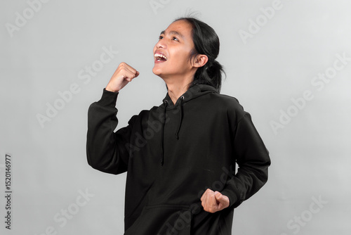 Asian man wearing a black hoodie expressing excitement and success with a triumphant fist gesture, looking up joyfully. Studio shot with neutral gray background.