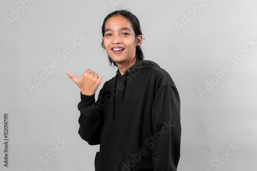 Portrait of a cheerful young Asian man with long hair wearing a black hoodie, pointing thumb to the side and smiling confidently against gray background.
