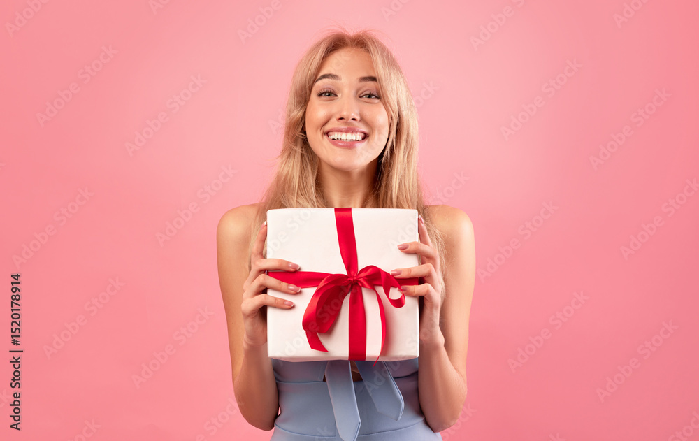 Fototapeta premium Spring holidays celebration. Cheerful millennial lady in pretty dress showing wrapped box with gift for Woman's Day or birthday on pink background. Young blonde feeling happy over special surprise