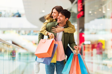 Couple Having Fun Shopping Together, Husband Carrying Wife Piggyback Posing With Colorful Paper Shopper Bags In Hypermarket. Black Friday Offer, Seasonal Sales Concept