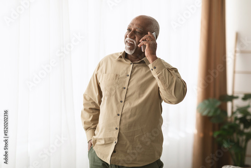 Mature Black Man Talking On Smartphone Looking Aside Standing Indoors Working From Home. Senior Male Having Pleasant Phone Conversation. Gadgets, Mobile Communication