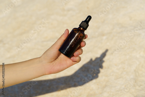 Female hand holding brown glass jar of serum with dispenser on stone background near the sea. Concept of natural beauty cosmetics, skin and face care.