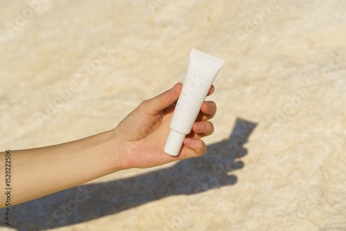 Female hand holding a white mockup tube of cream or serum on a stone background illuminated by the sun. Concept of cosmetic products, skin hydration and nourishment, anti-aging, smoothing.