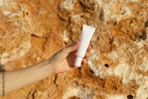 Close-up of a female hand holding a thin white mockup tube of eye cream or serum on a limestone background near the sea. Concept of skin care, moisturizing, nutrition, sunscreen, anti-aging.