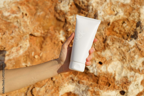 Female hand holding white mockup tube of cream on limestone background near the sea. Concept of skin care, moisturizing, beauty products, sunscreen, sun and ultraviolet protection with SPF.