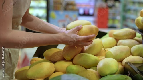 Tourist woman examing mango fruit, squeezing to test ripeness before buy. Fresh tropical fruit selection, grocery shopping, travel destination in grocery market, local produce quality checked
