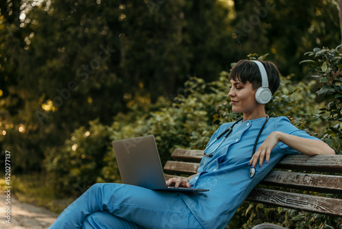 Doctor wearing headphones and using laptop while relaxing on bench in park