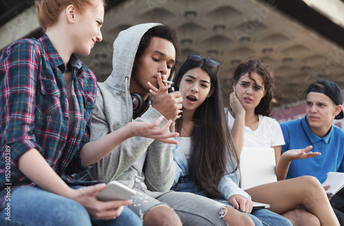 Obraz na plátne Black teen guy smoking cigarette, his friends judging him