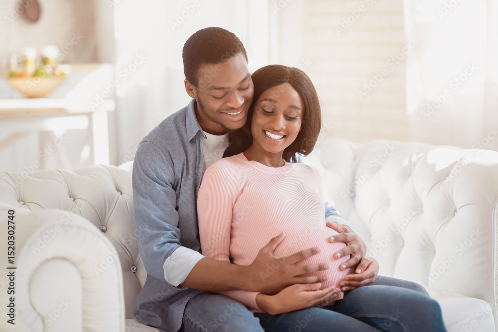 Smiling future mom and dad expecting baby, sitting on sofa at home and hugging. Beautiful pregnant couple embracing on couch. Young African American guy embracing his expectant wife Smiling future mom and dad expecting baby, sitting on sofa at home and hugging. Beautiful pregnant couple embracing on couch. Young African American guy embracing his expectant wife